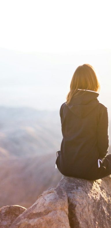 Medical Coach | Physiotherapie München & Stanzach Eine Person sitzt auf einem Felsen und blickt auf eine bergige Landschaft bei Sonnenlicht.