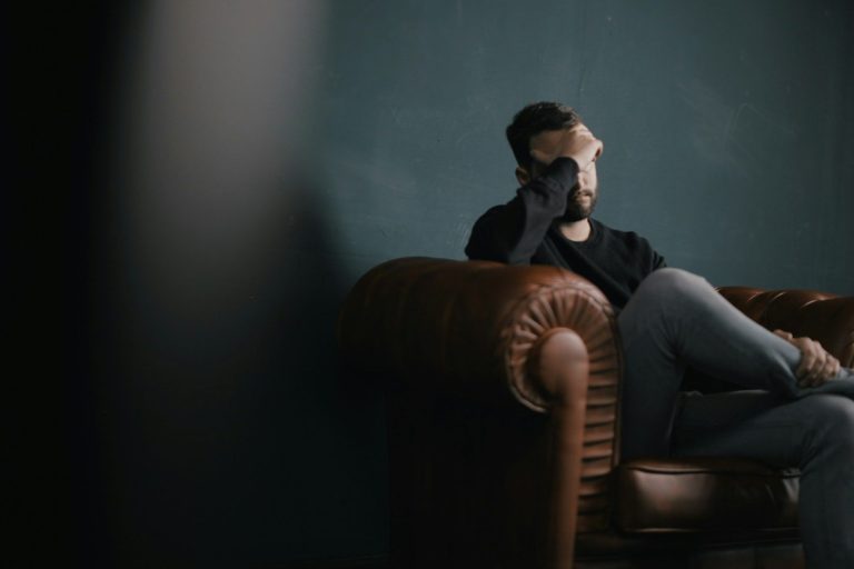 Schmerztherapie München & Stanzach Mann sitzt nachdenklich auf einem Sofa mit verschränkten Beinen.